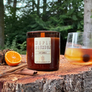 Rustic Maple Bourbon Soy Candle in amber tumbler jar with wood wick on tree stump in forest with slice orange and Old Fashioned cocktail pine cone cinnamon sticks